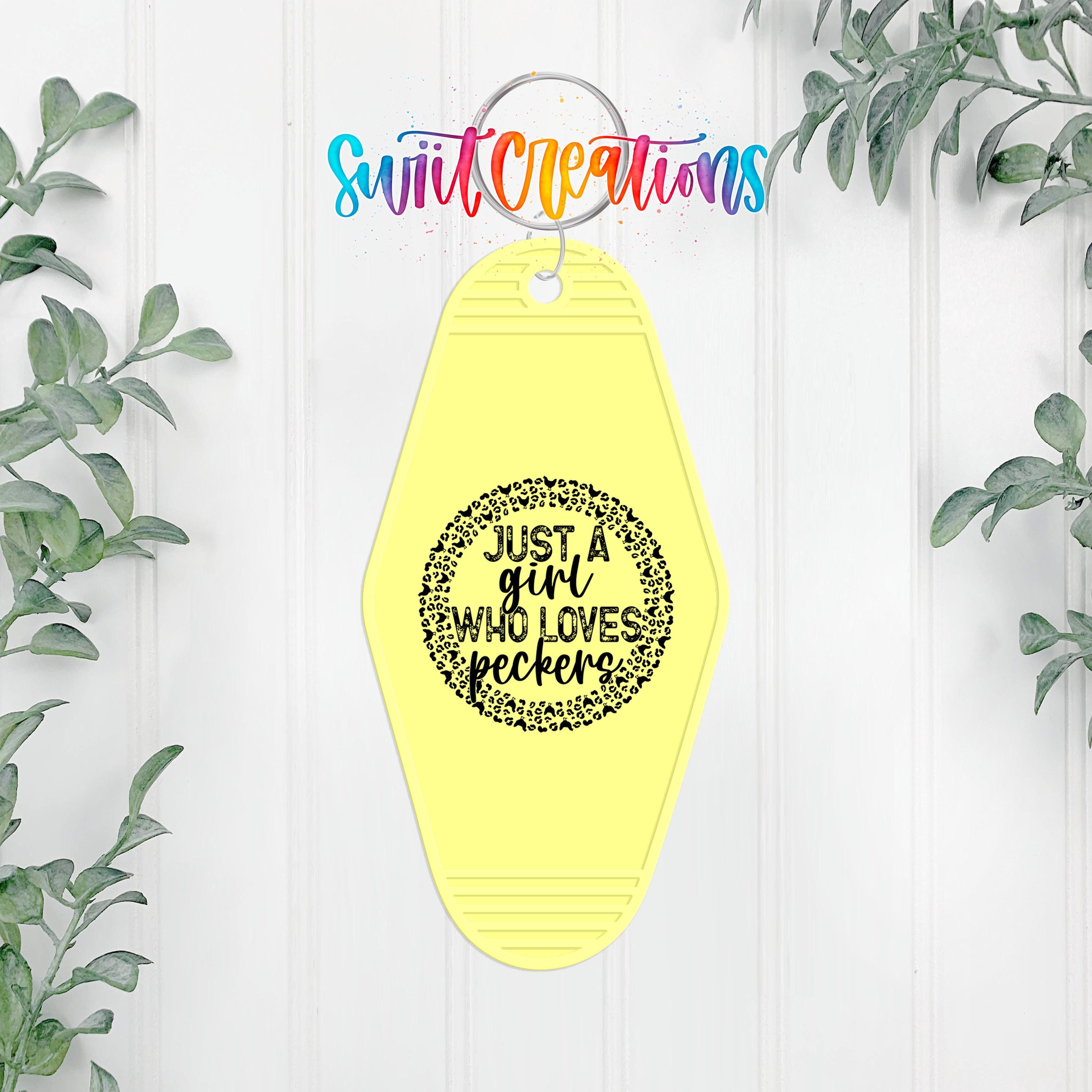 A yellow keychain with a black text design that says "Just a girl who loves peckers" is hanging on a white wall, surrounded by greenery.