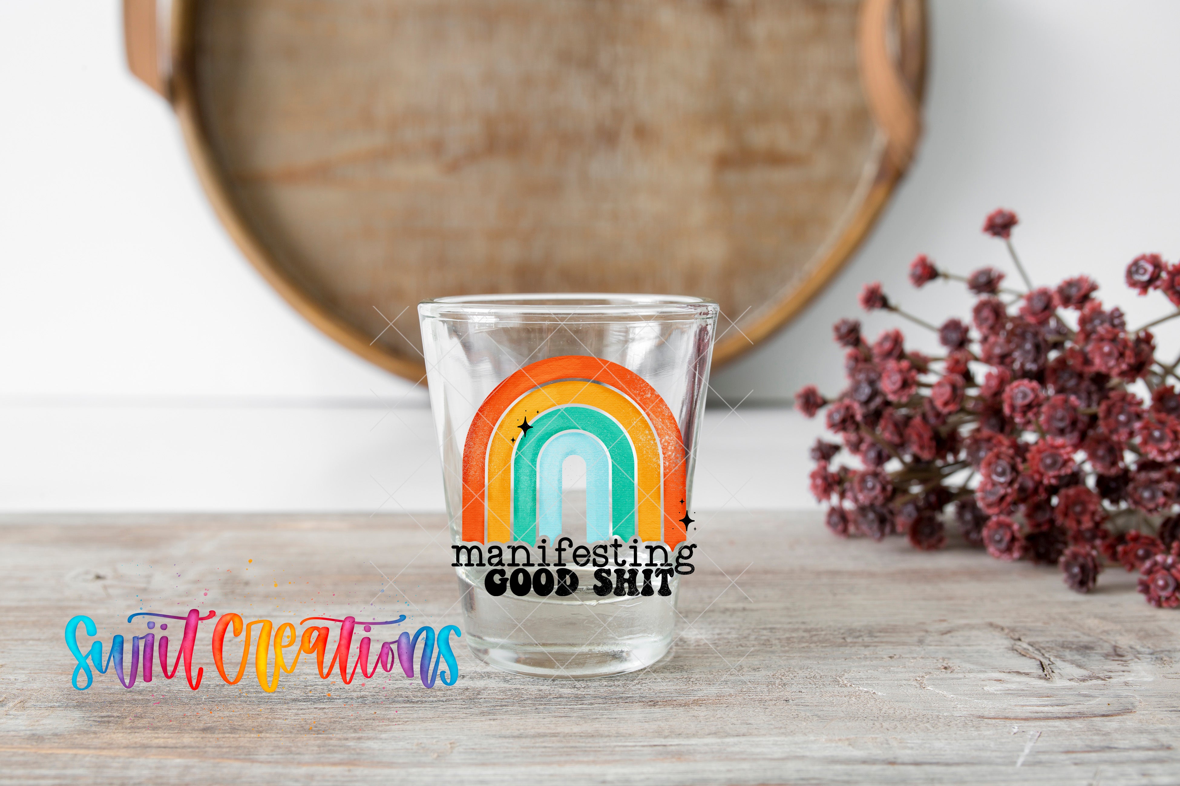 A glass with a rainbow-colored design and the text "manifesting good shit" is placed on a wooden surface, with a bunch of flowers and a wooden tray in the background.