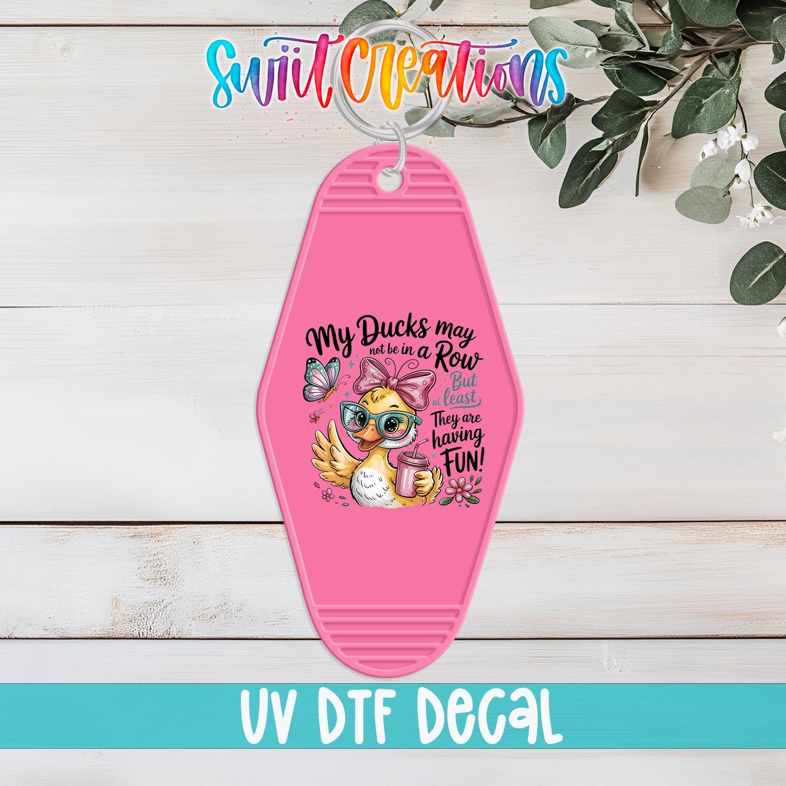 A pink keychain with a cartoon duck and text, placed on a wooden surface with greenery in the background.