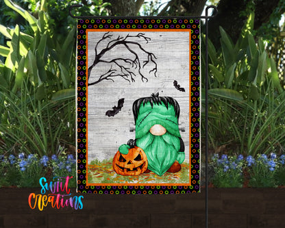 A Halloween-themed garden flag featuring a green gnome holding a carved pumpkin, with a spooky tree and bats in the background.