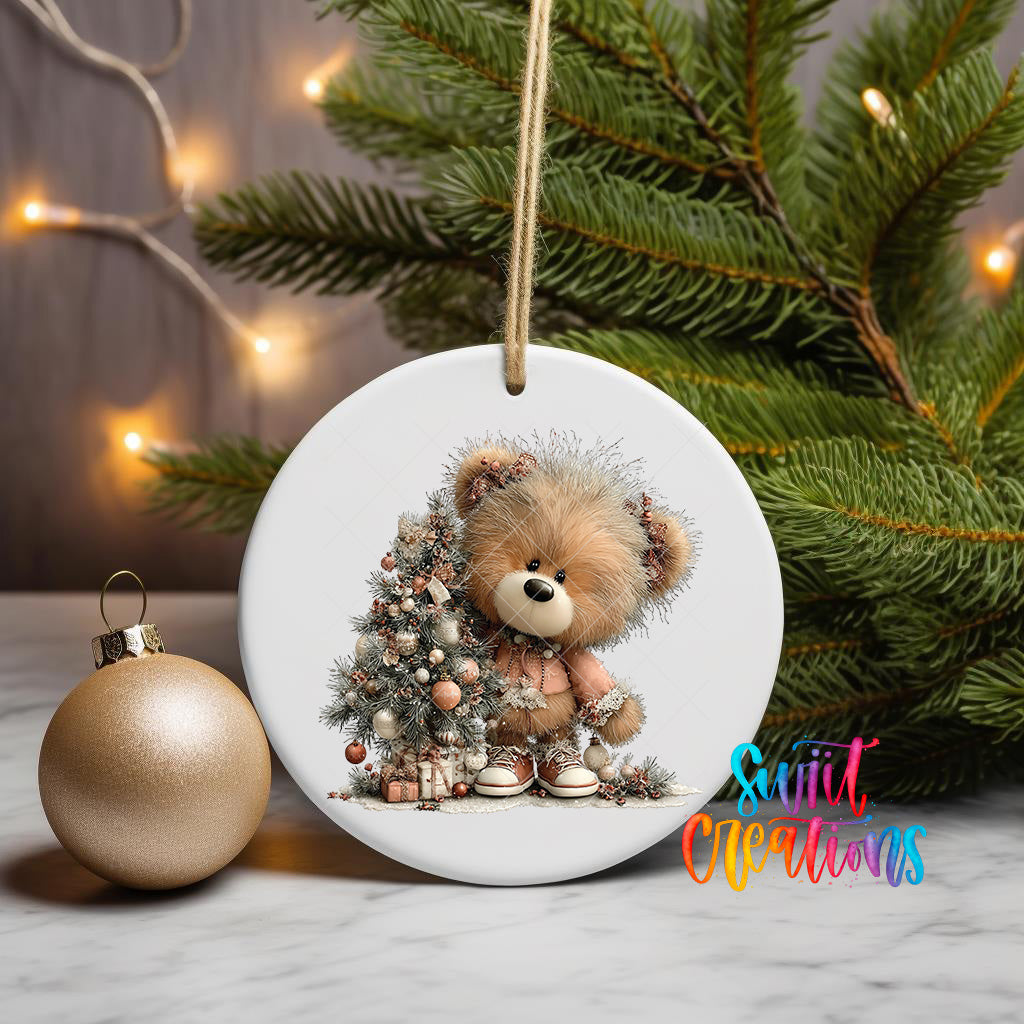 A white ceramic ornament featuring a teddy bear sitting in front of a Christmas tree, with a gold ornament to the left and a blurred background of greenery and twinkling lights.