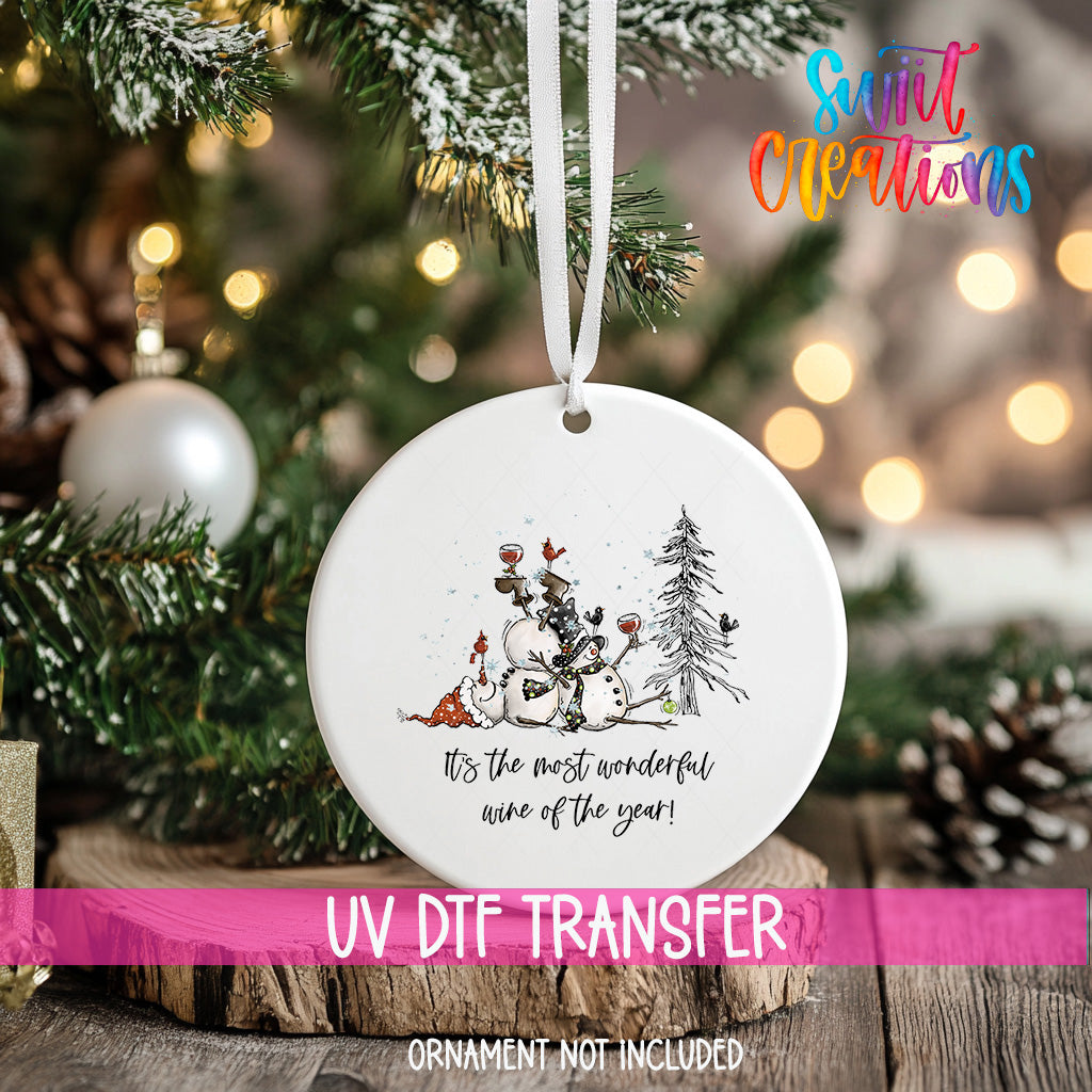 A white ornament with a snowman and Christmas tree design hangs on a Christmas tree, with a pink ribbon around it.