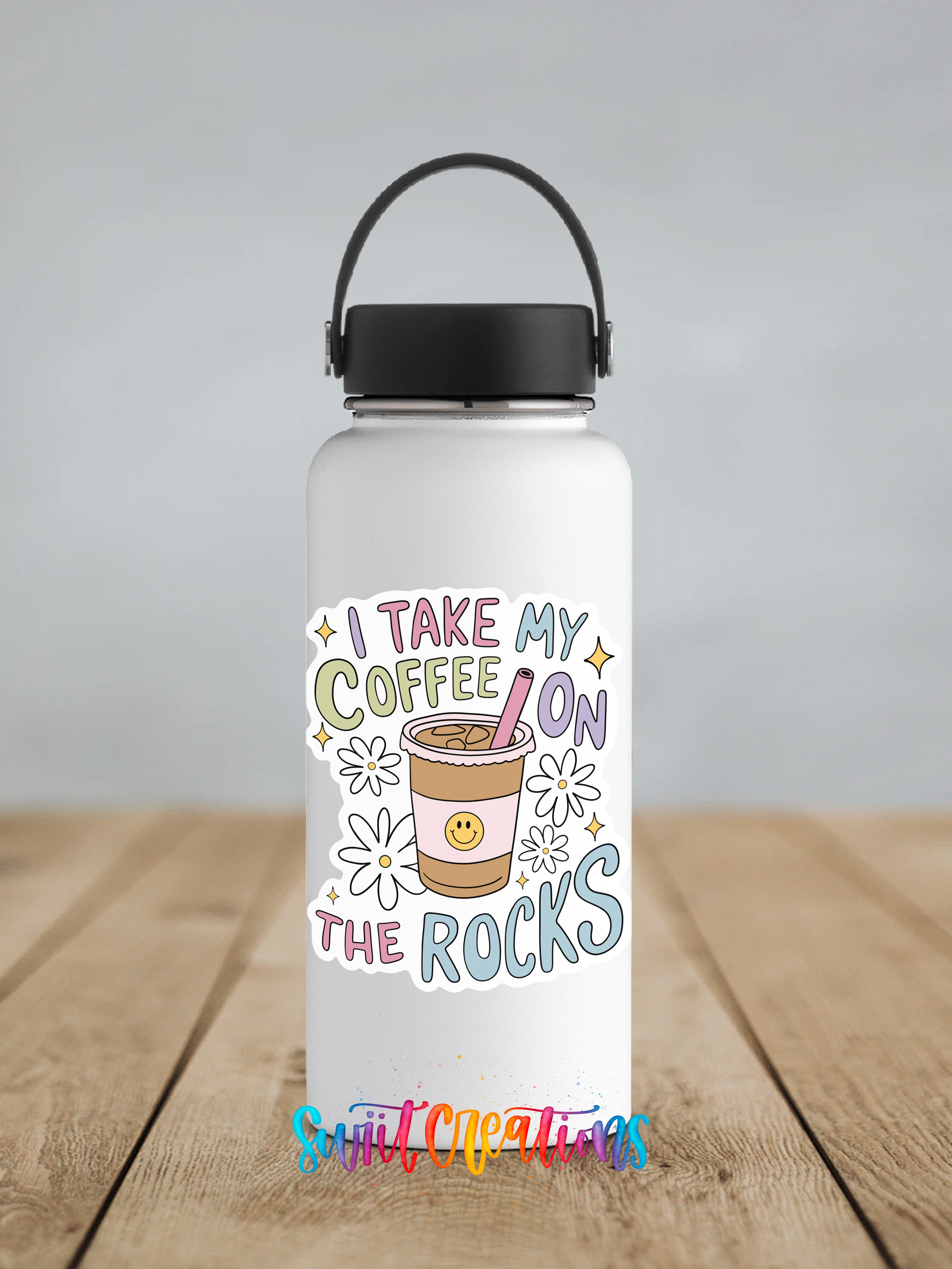 A white water bottle with a colorful sticker that says "I TAKE MY COFFEE ON THE ROCKS" and features a coffee cup and flowers.