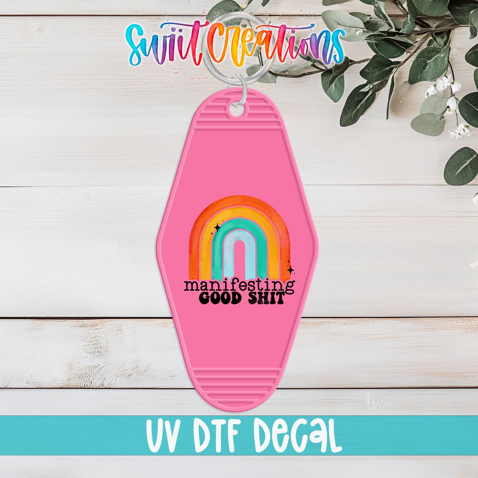 A pink keychain with a rainbow graphic and the text "manifesting good shit" printed on it, placed on a wooden surface with some greenery in the background.