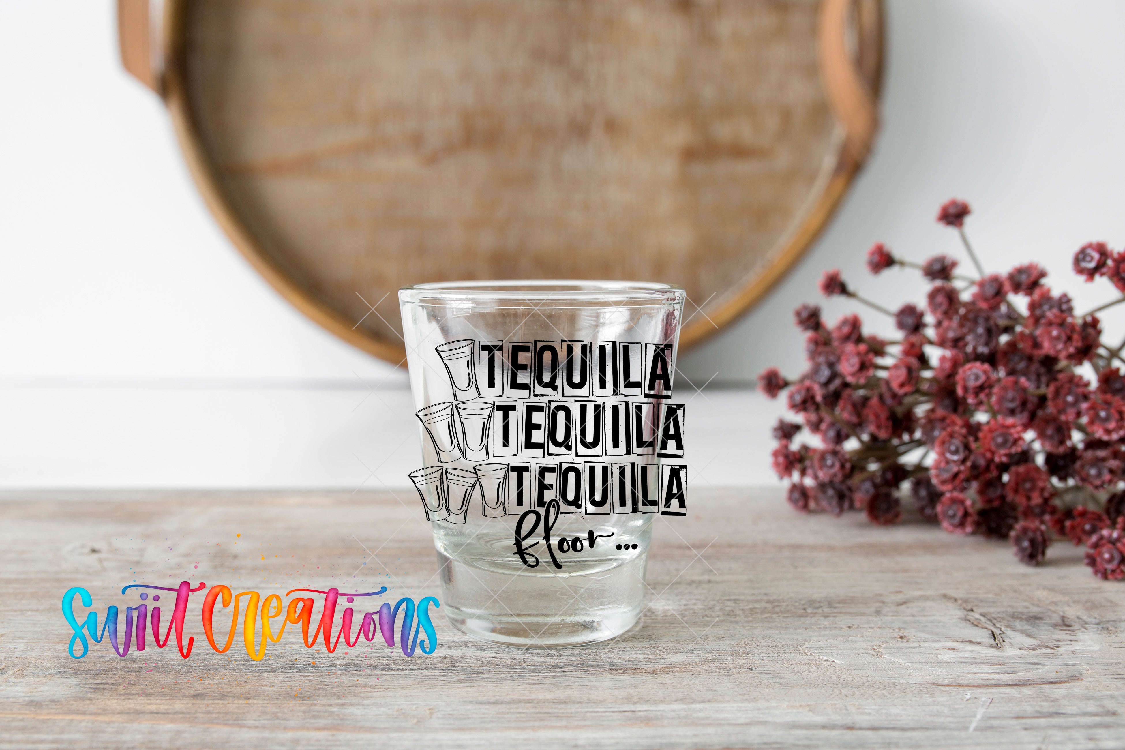 A clear glass with the text "TEQUILA TEQUILA TEQUILA" printed on it, along with a colorful graphic of tequila glasses, sits on a wooden surface. In the background, there is a wooden tray and some flowers.