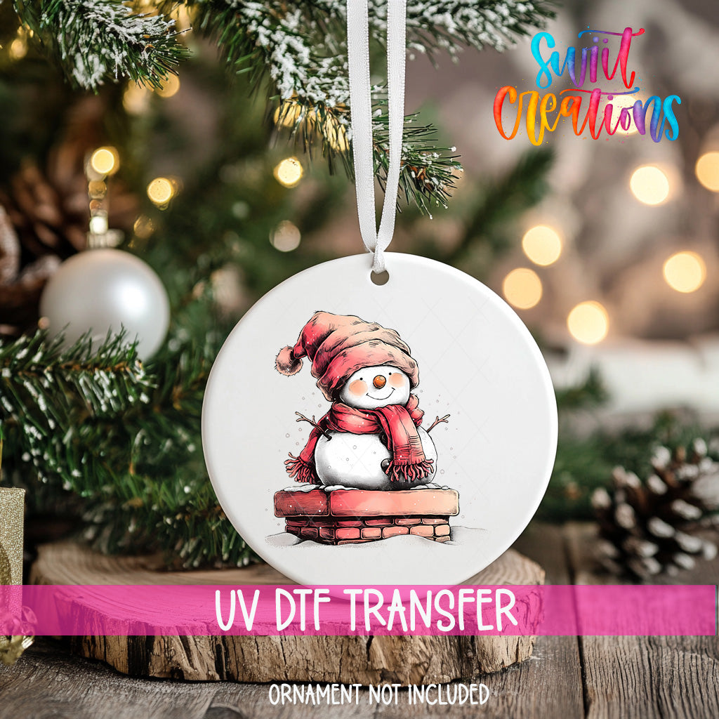 A white ornament with a snowman design hangs on a Christmas tree, surrounded by pine branches and twinkling lights.