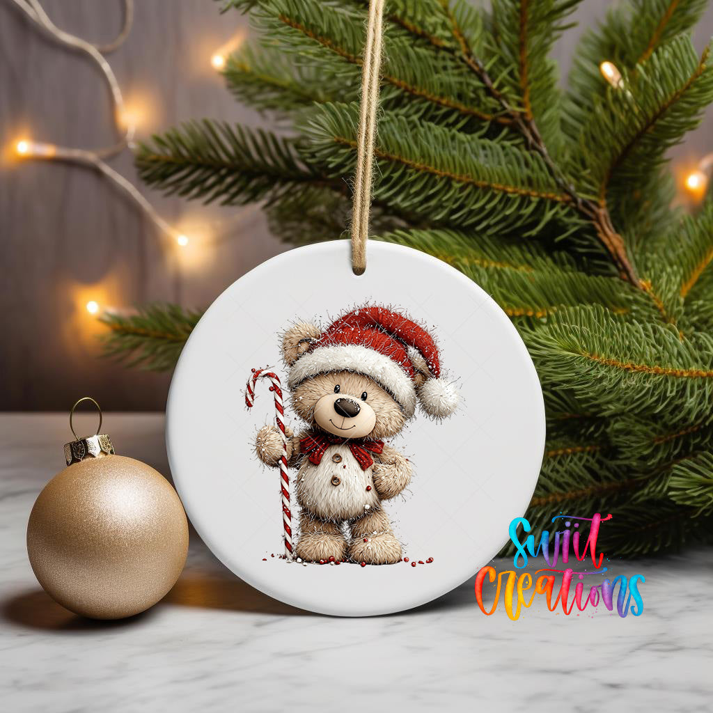 A white ceramic ornament featuring a teddy bear wearing a Santa hat and holding a candy cane, placed on a marble surface next to a gold ornament and a Christmas tree.