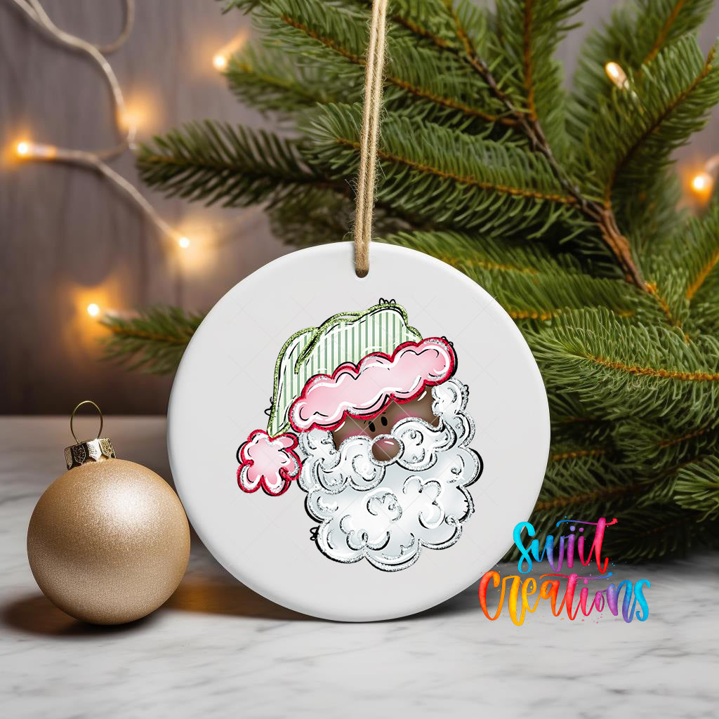 A white ceramic ornament with a Santa Claus face design is displayed on a white surface, accompanied by a gold ornament and a Christmas tree with twinkling lights in the background.