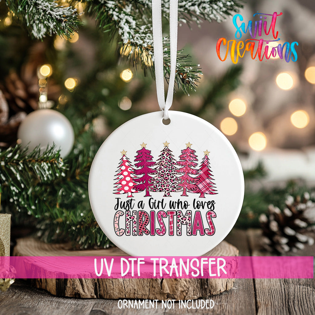 A white Christmas ornament with a design of pink and white trees hangs from a tree branch, with a blurred background of twinkling lights.