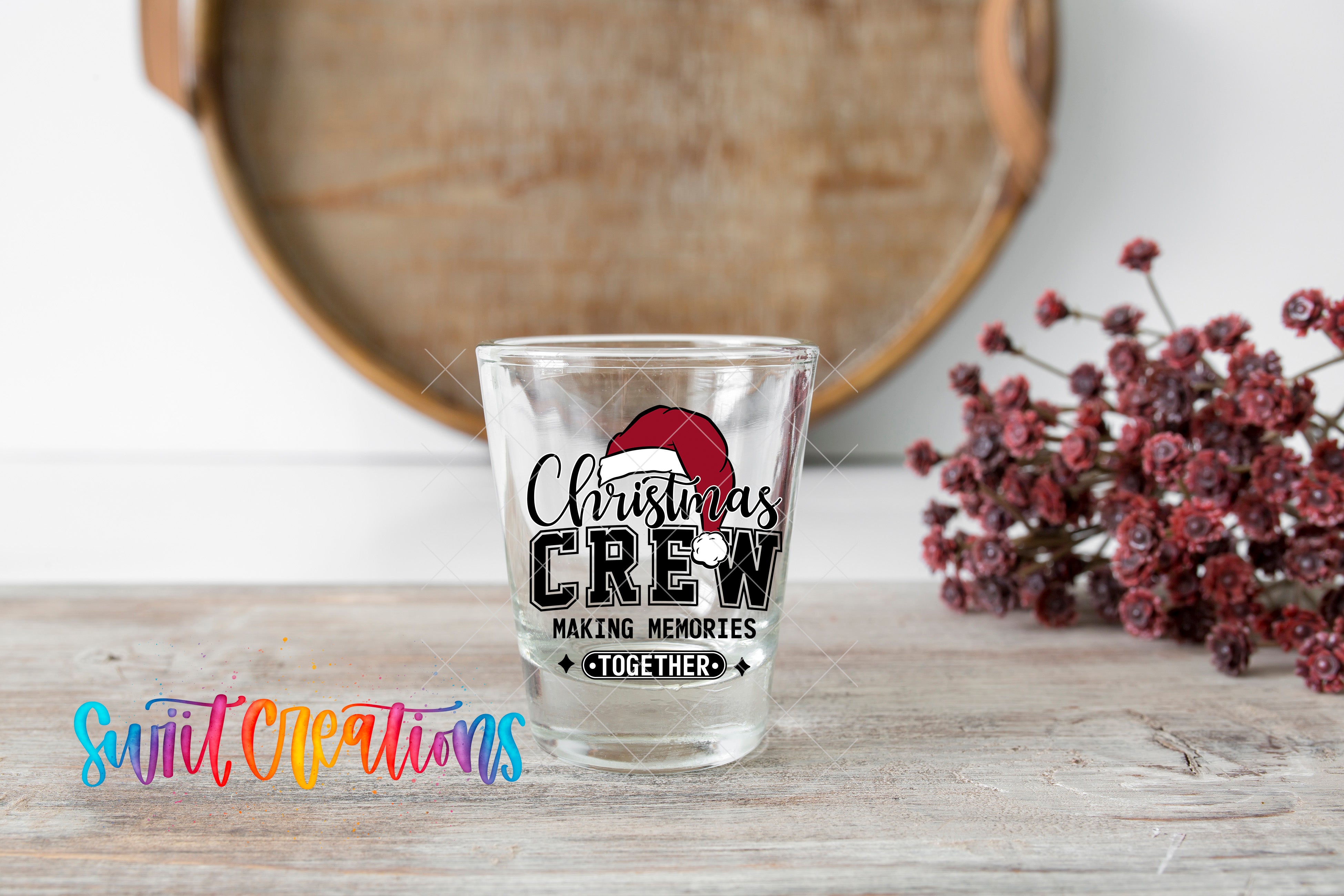 A shot glass with a Christmas crew design on it, placed on a wooden surface next to a bunch of red flowers.