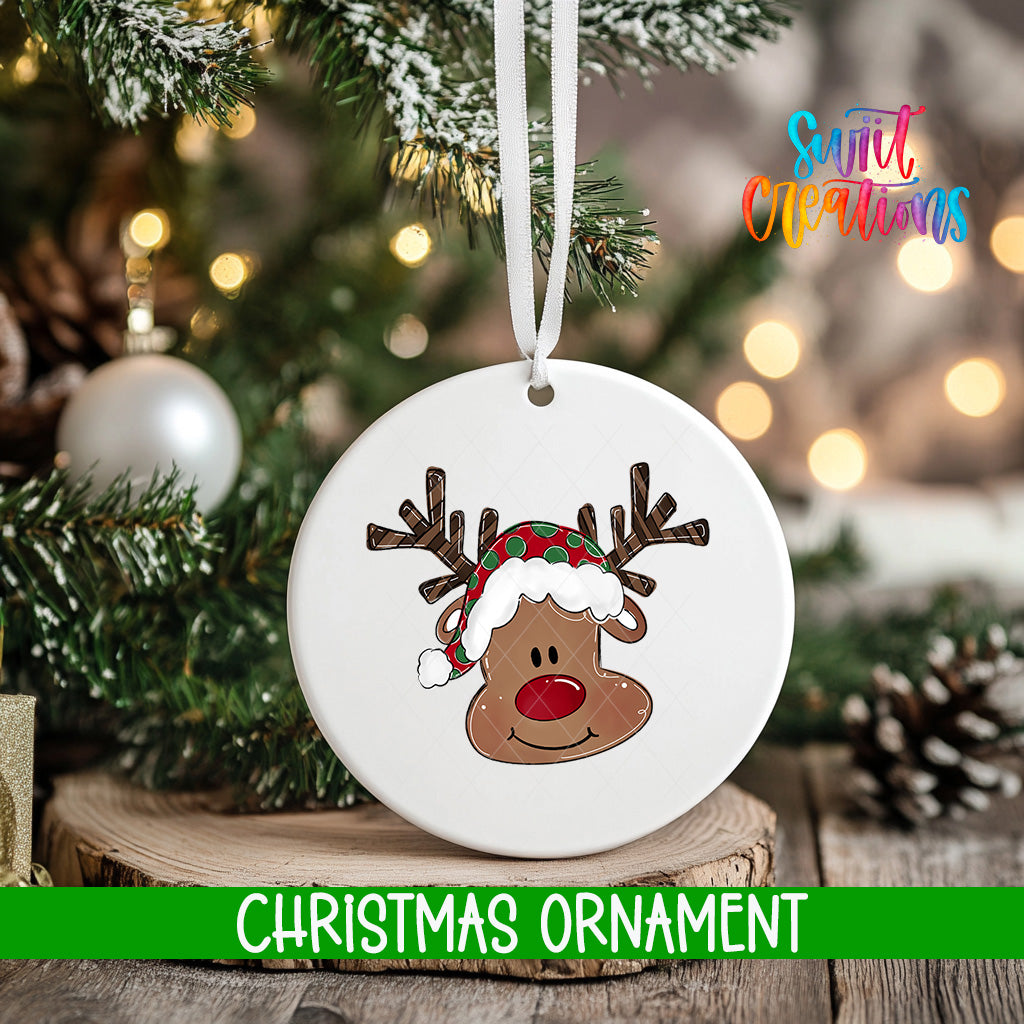 A white Christmas ornament with a cartoon reindeer wearing a Santa hat is hanging on a tree branch.