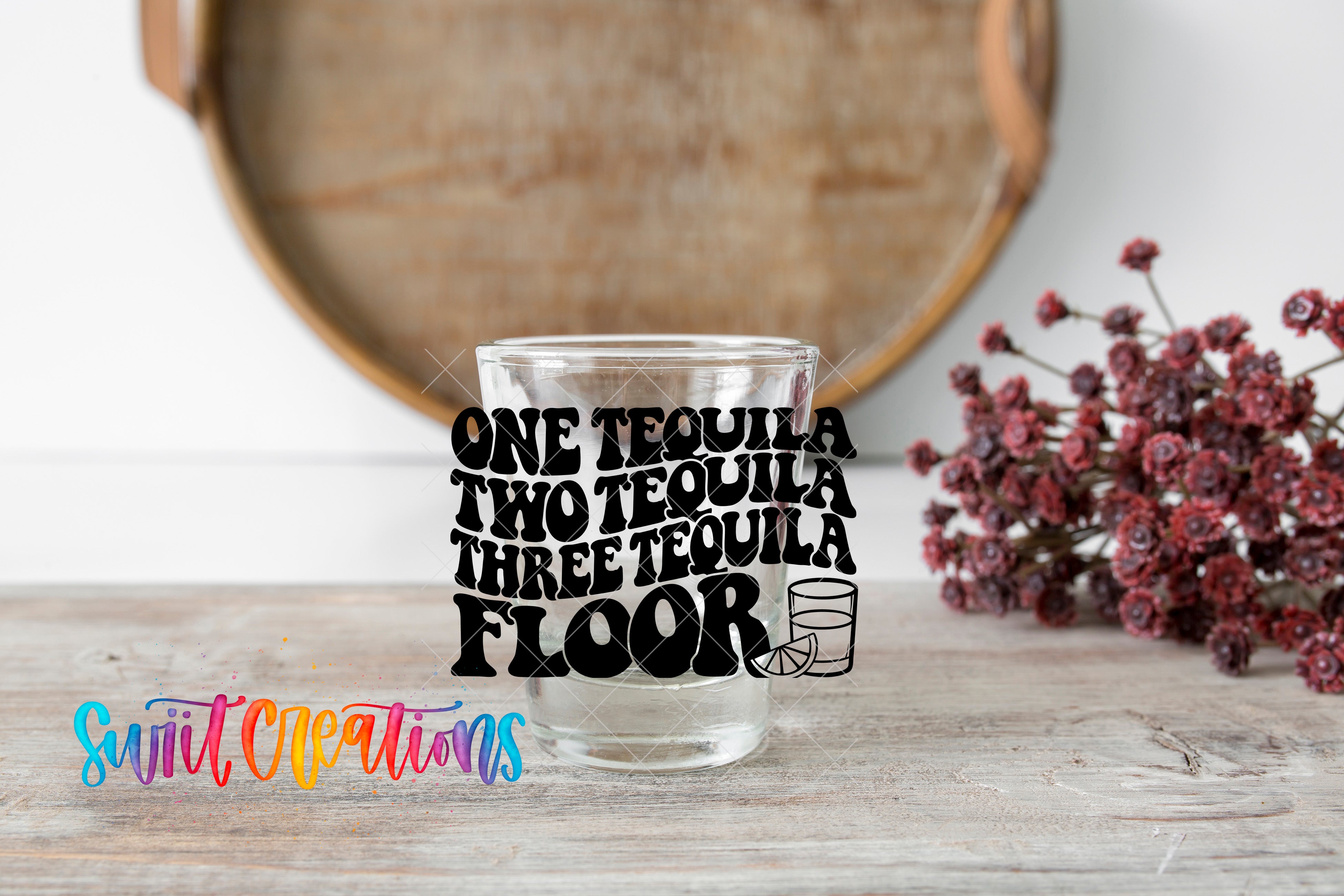 a clear glass with the text "ONE TEQUILA TWO TEQUILA THREE TEQUILA FLOOR" printed on it, along with a graphic of a tequila glass and a lime. The glass is placed on a wooden surface, and there is a bouquet of flowers in the background.
