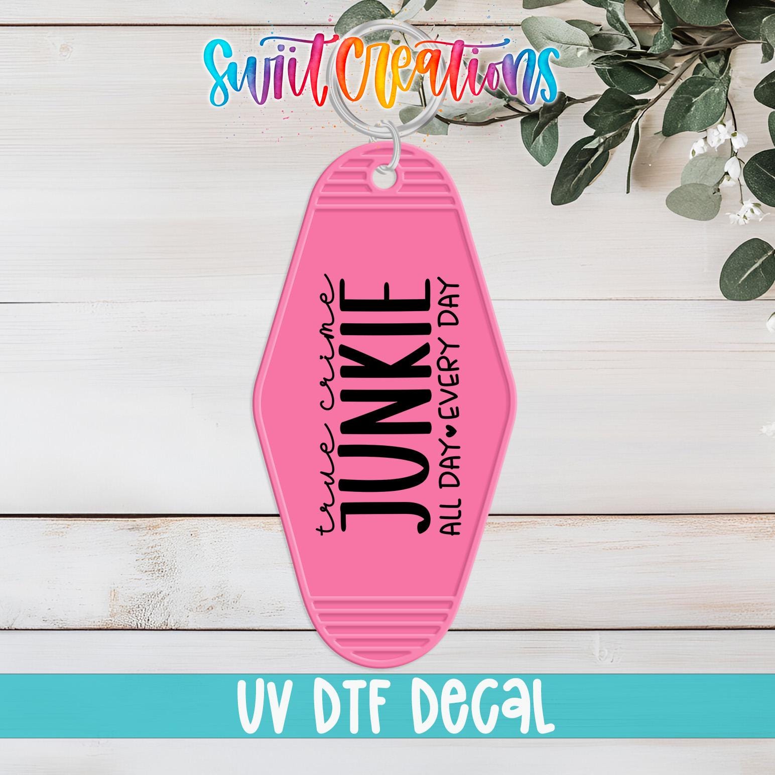 A pink keychain with the text "True Crime Junkie All Day" printed on it, placed on a wooden surface with some greenery in the background.