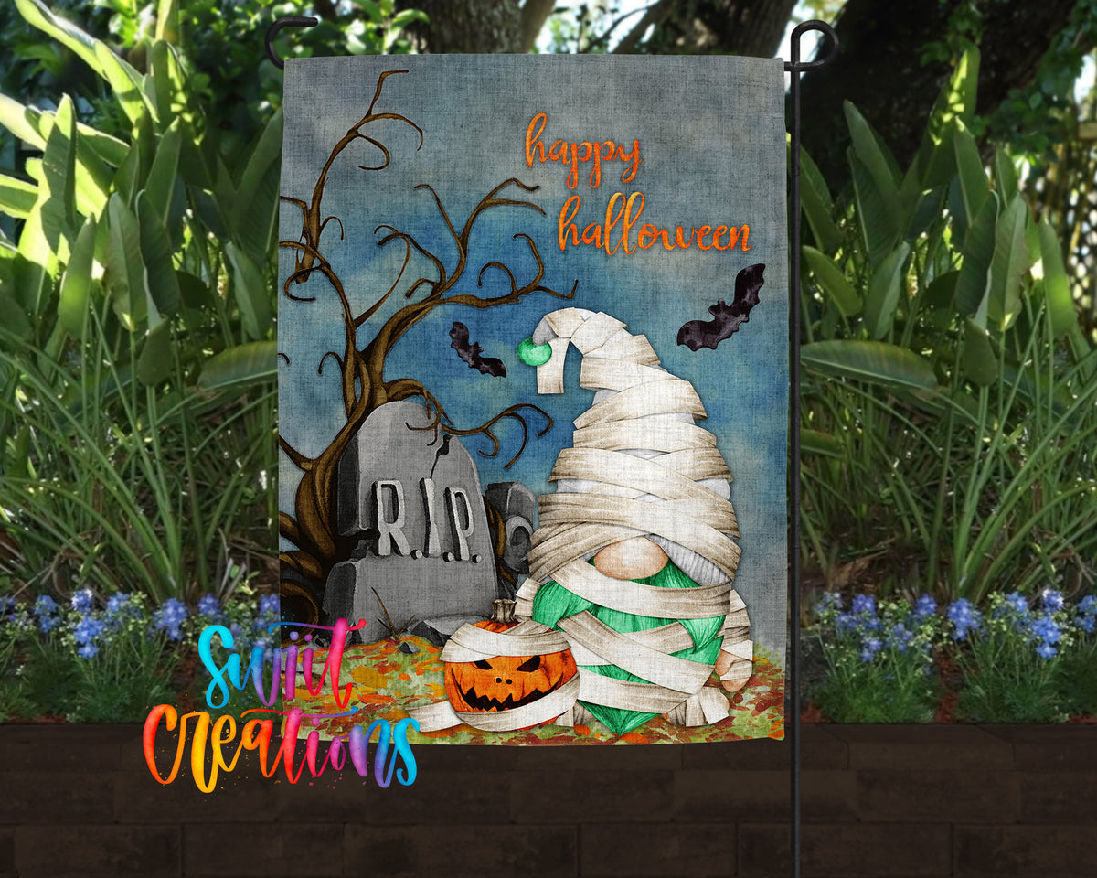 A Halloween-themed garden flag featuring a gnome wrapped in white cloth, a carved pumpkin, a gravestone, and bats flying overhead.