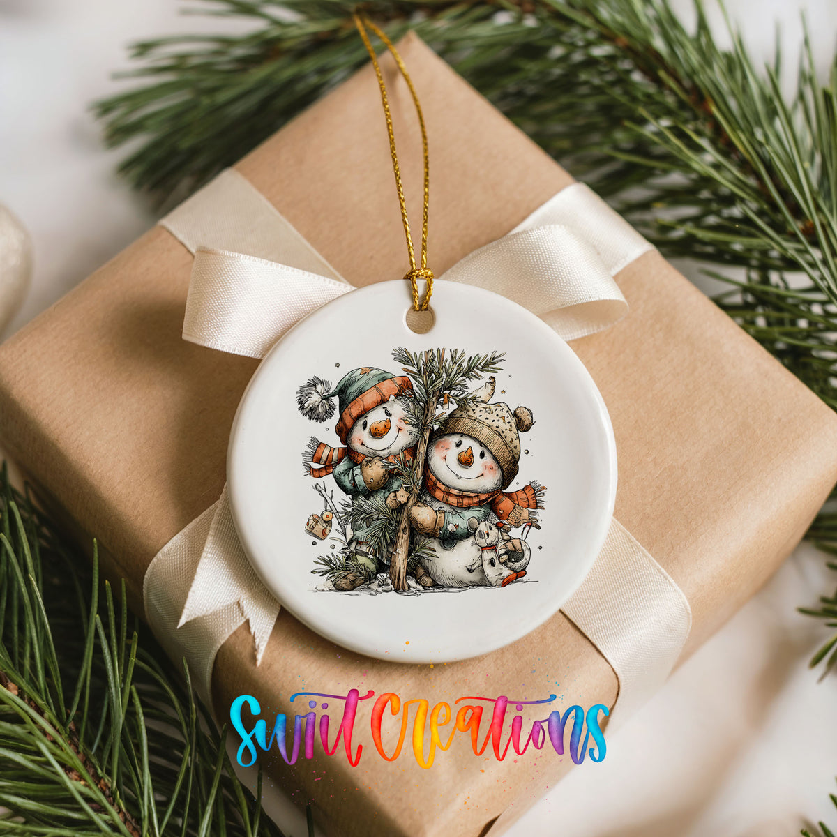 A white ceramic ornament with a snowman and pine tree design, hanging from a ribbon. The ornament is placed on top of a brown gift box, surrounded by pine branches.