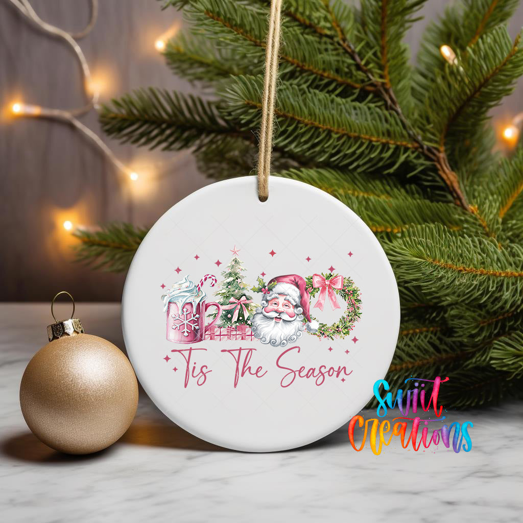 A white ceramic ornament with a Santa Claus and Christmas tree design, along with the text "Tis the Season" and a small gold ornament, is displayed on a marble surface.