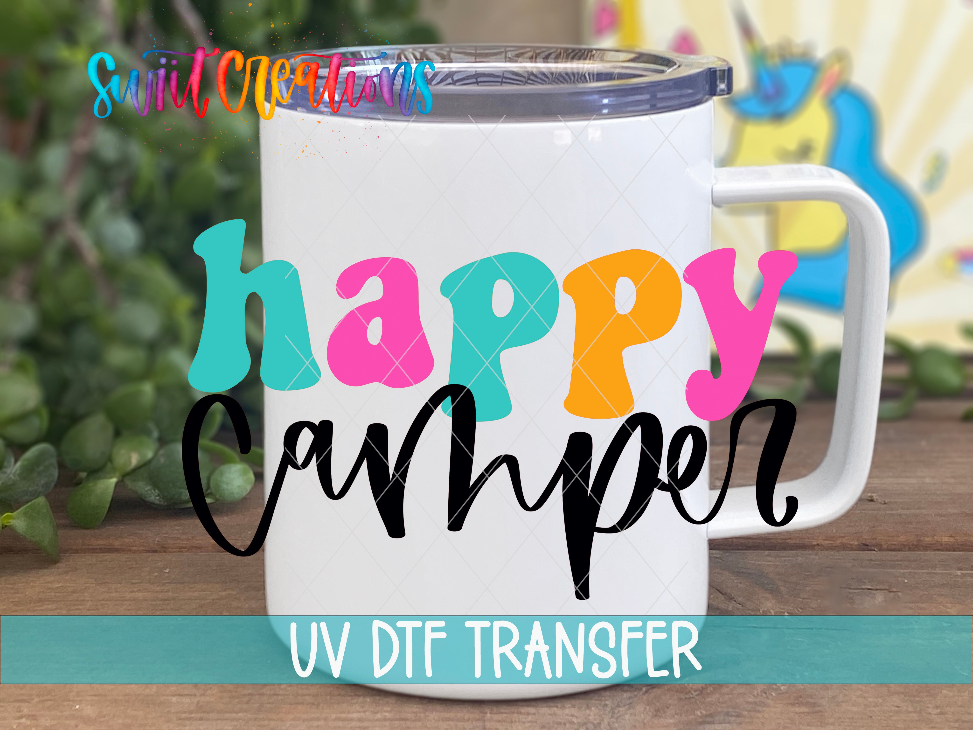 A white ceramic mug with colorful text that says "hAPPY Camper" on it, placed on a wooden surface with a blurred background.