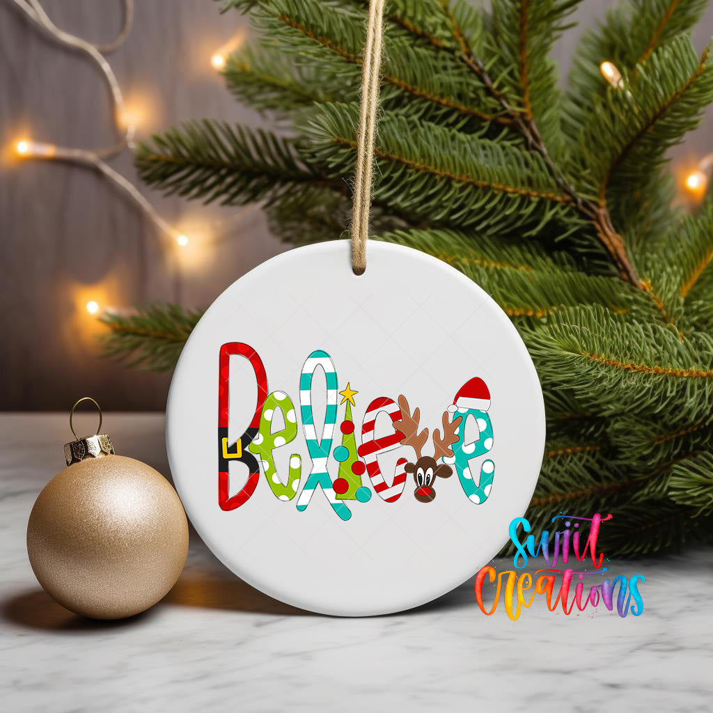 A white ceramic ornament with the word "Believe" written on it, hanging from a string, with a gold ornament and a pine tree in the background.