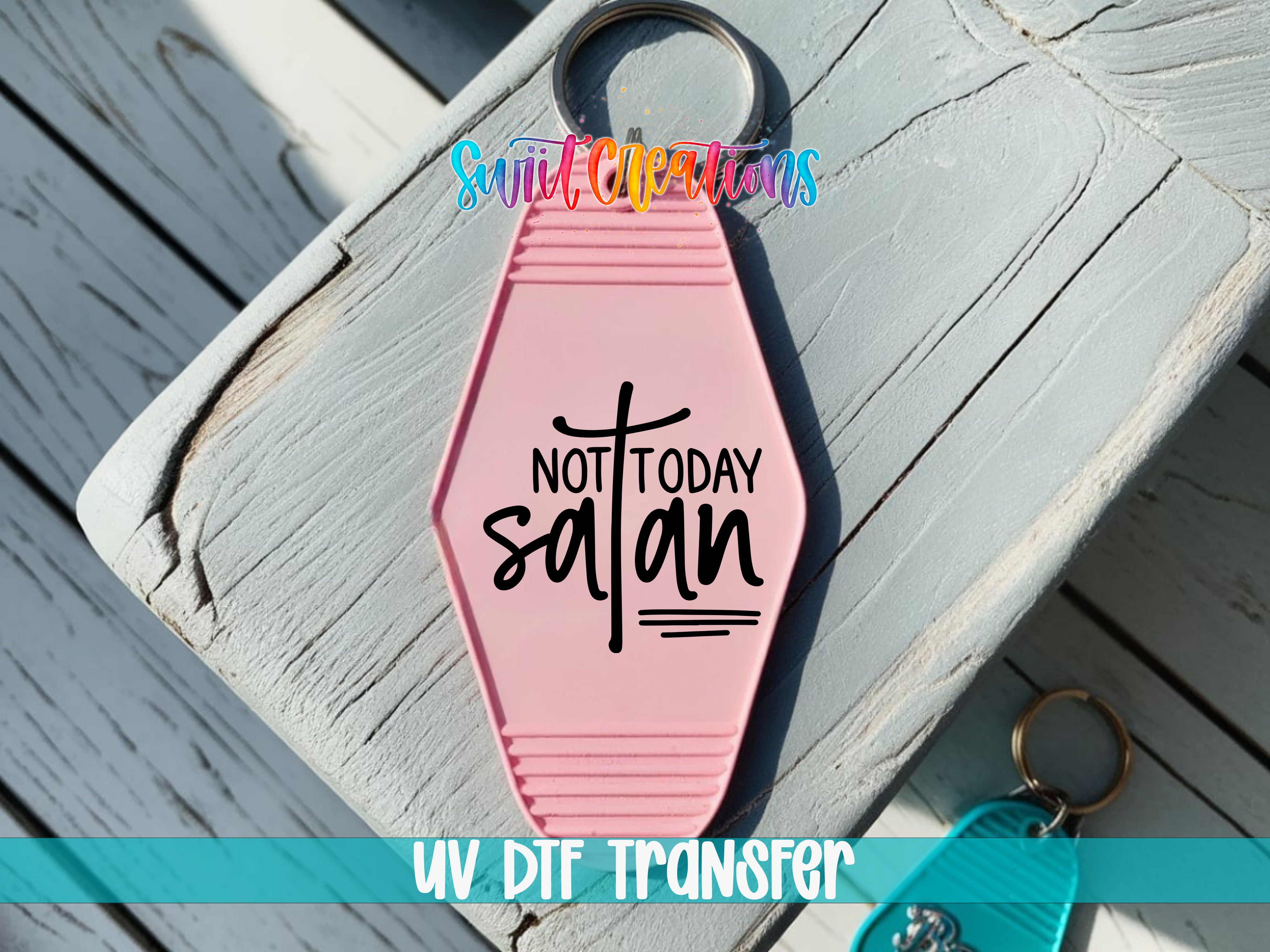 A pink keychain with the text "NOT TODAY SATAN" printed on it, placed on a wooden surface.