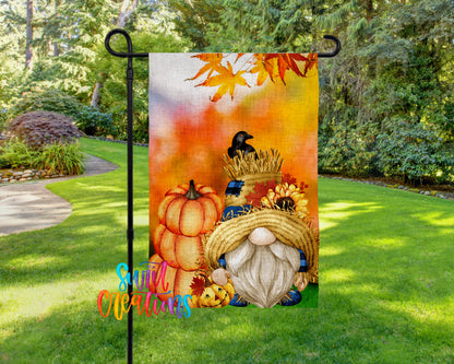 A colorful autumn-themed garden gnome flag is displayed on a metal pole in a lush, green garden setting.