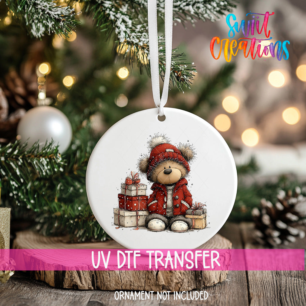 A white ornament with a teddy bear and presents is hanging on a Christmas tree, with a pink ribbon around it.