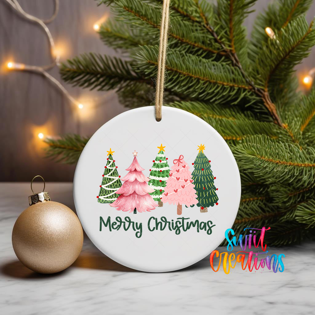 A white ceramic ornament with a festive design of Christmas trees and a gold ornament is displayed on a marble surface, with a string of lights and a pine tree in the background.