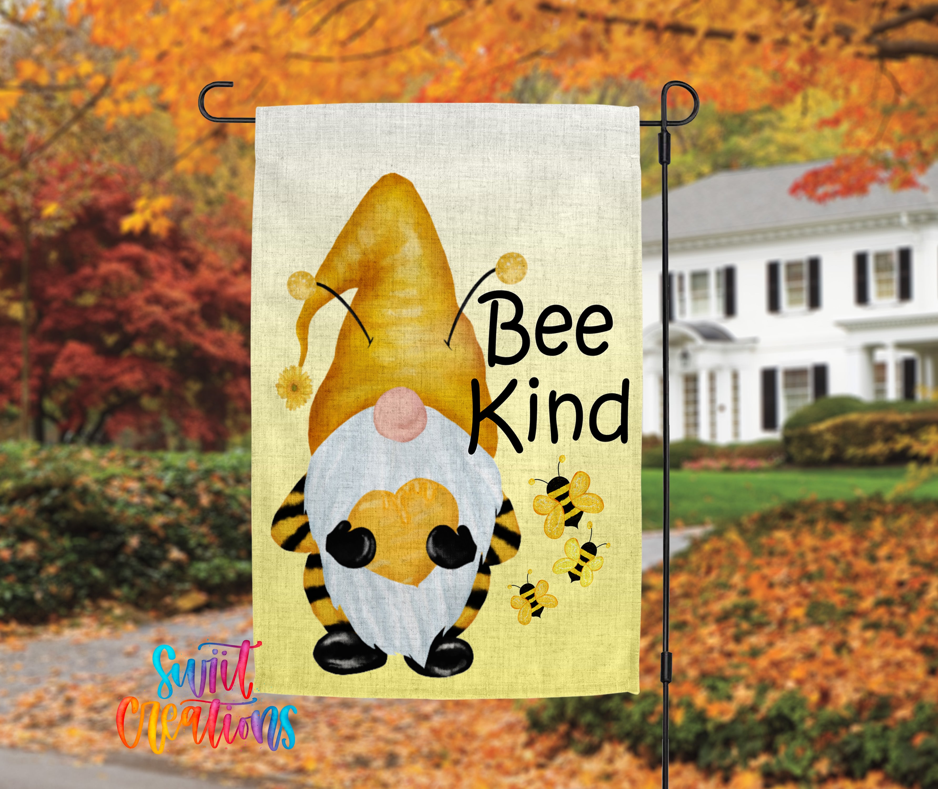 A garden gnome with a yellow hat and a sign that says "Bee Kind" is displayed in the foreground, with a white house and colorful autumn foliage in the background.