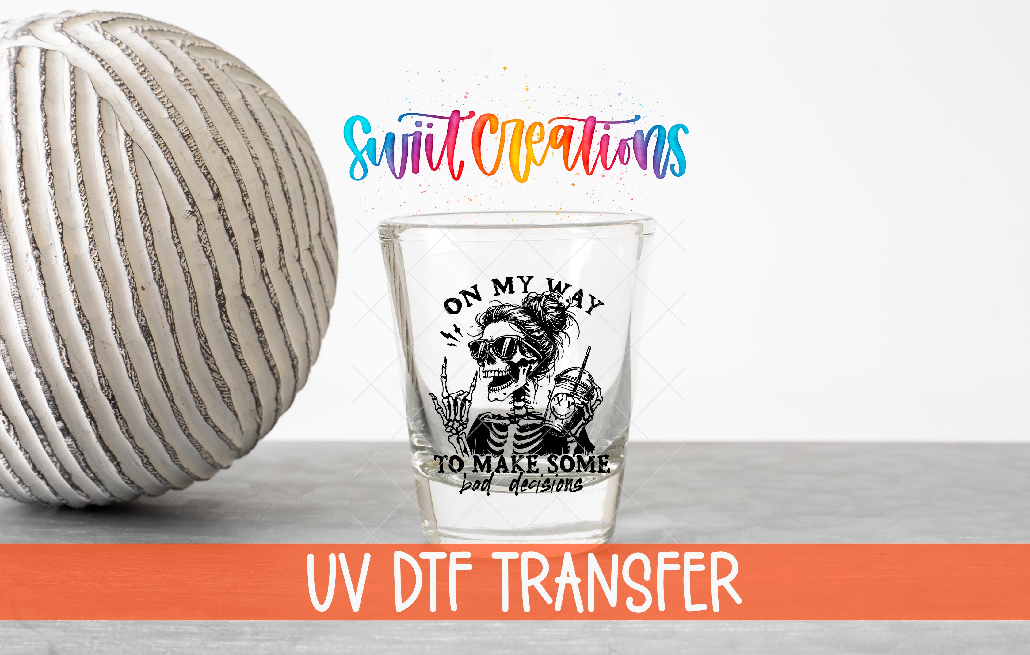 a shot glass with a skull design and text, placed next to a striped ball and a text overlay that says "Swit Creations UV Dtf Transfer".