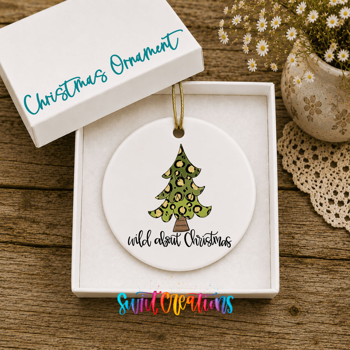A white Christmas ornament with a leopard print design is displayed in a white box, accompanied by a small potted plant and a lace doily on a wooden surface.