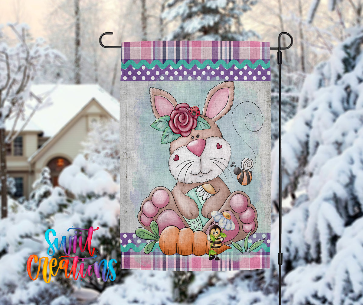 A colorful garden flag featuring a cartoon rabbit holding a flower and a bee, set against a snowy background with a house and trees in the distance.