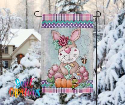 A colorful garden flag featuring a cartoon rabbit holding a flower and a bee, set against a snowy background with a house and trees in the distance.