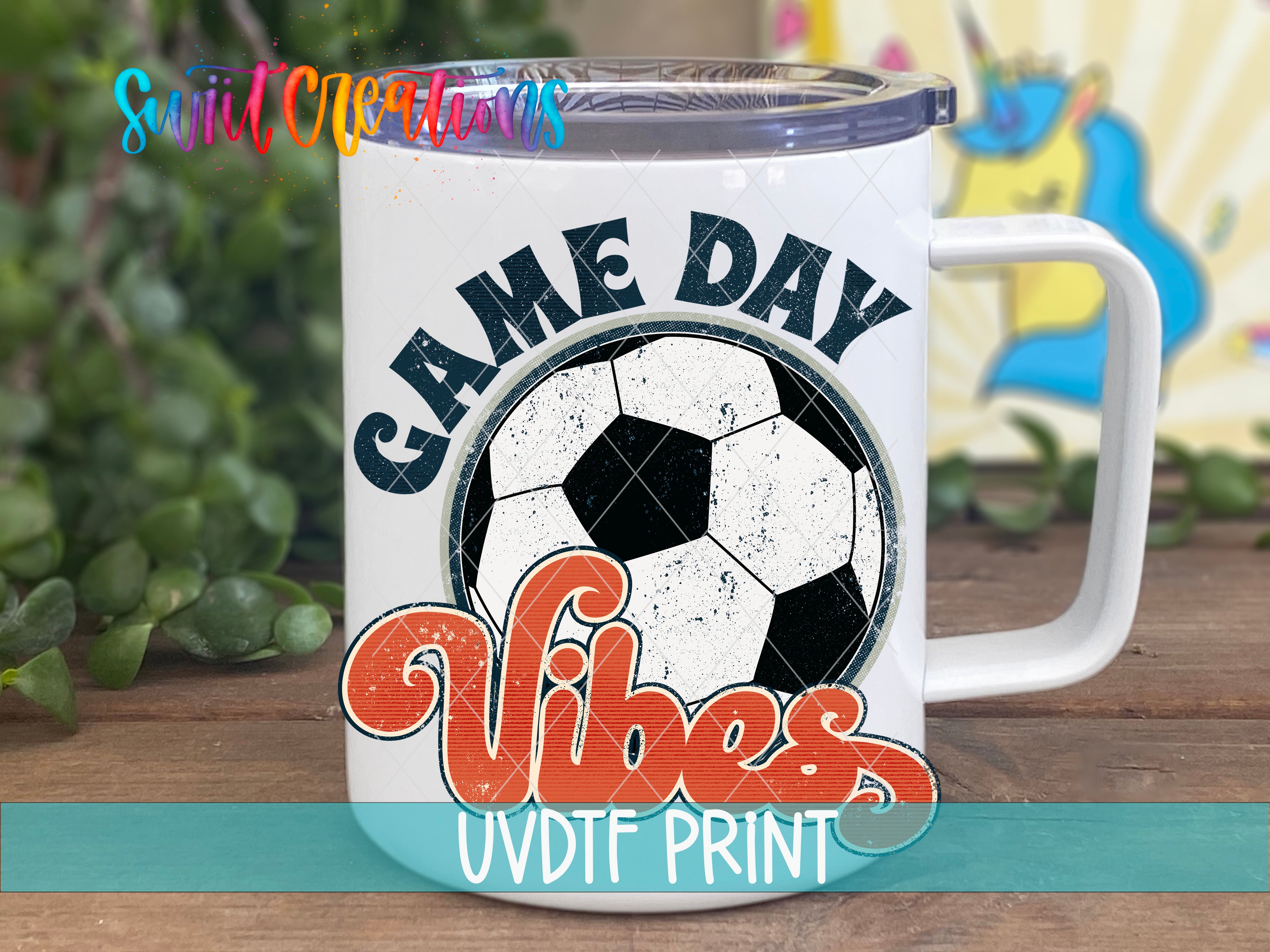 A white mug with a soccer ball design and the text "GAME DAY VIBES" printed on it, placed on a wooden surface.