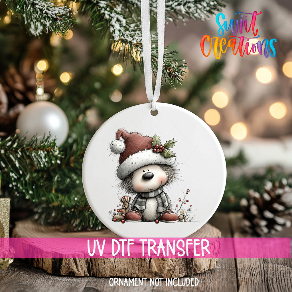 A white ornament with a cartoon image of a dog wearing a Santa hat and scarf is hanging on a Christmas tree.