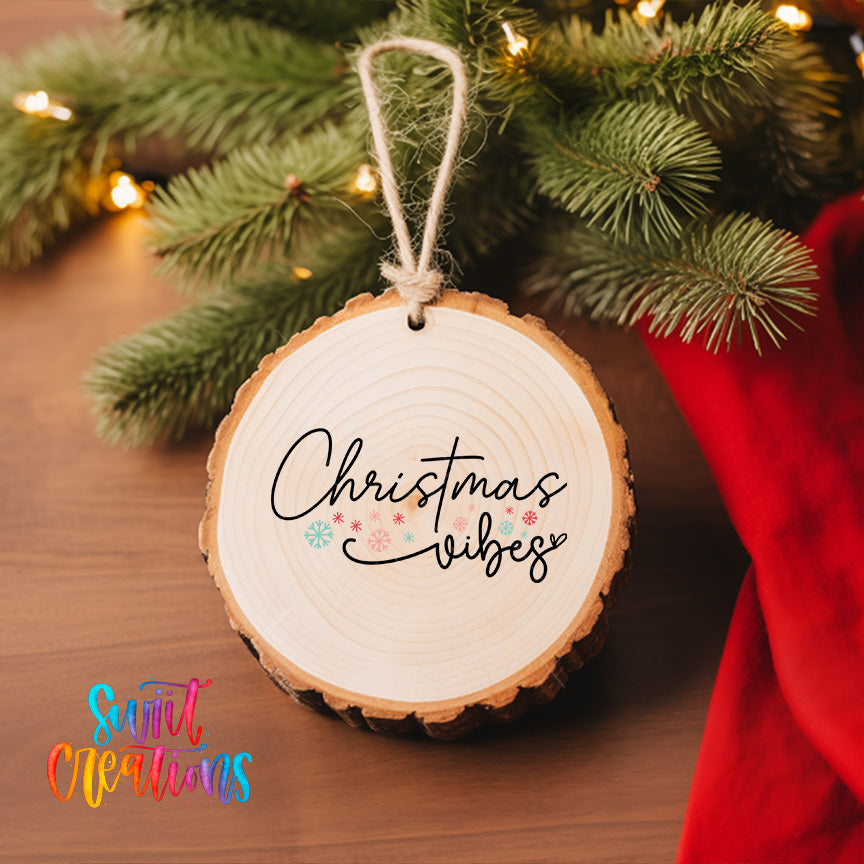 A wooden ornament with the text "Christmas vibes" hanging from a tree branch, surrounded by pine branches and twinkling lights.