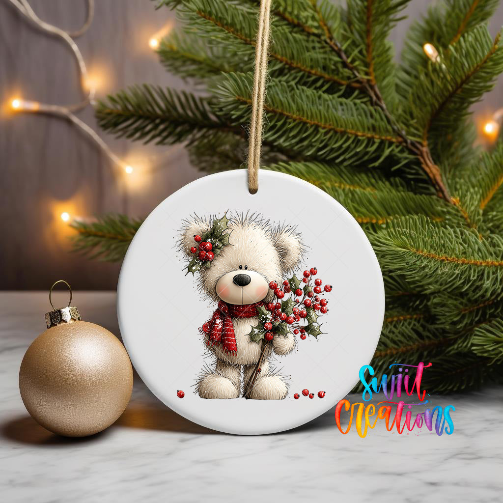 A white ceramic ornament featuring a teddy bear holding berries, placed on a marble surface next to a gold ornament and a Christmas tree with twinkling lights.
