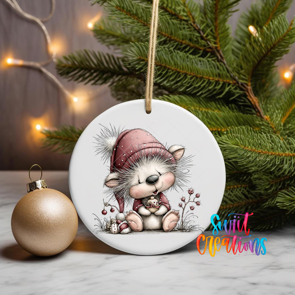 A ceramic ornament featuring a smiling gnome wearing a red hat and scarf, sitting on a branch with pine needles and berries, is displayed on a white surface next to a gold ornament and a Christmas tree.