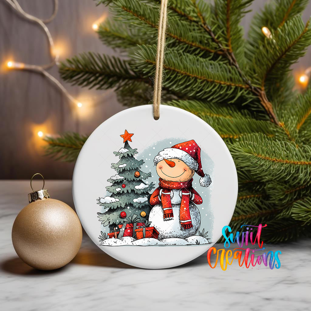A white ceramic ornament featuring a snowman and a Christmas tree, with a star on top, is displayed on a table next to a gold ornament.