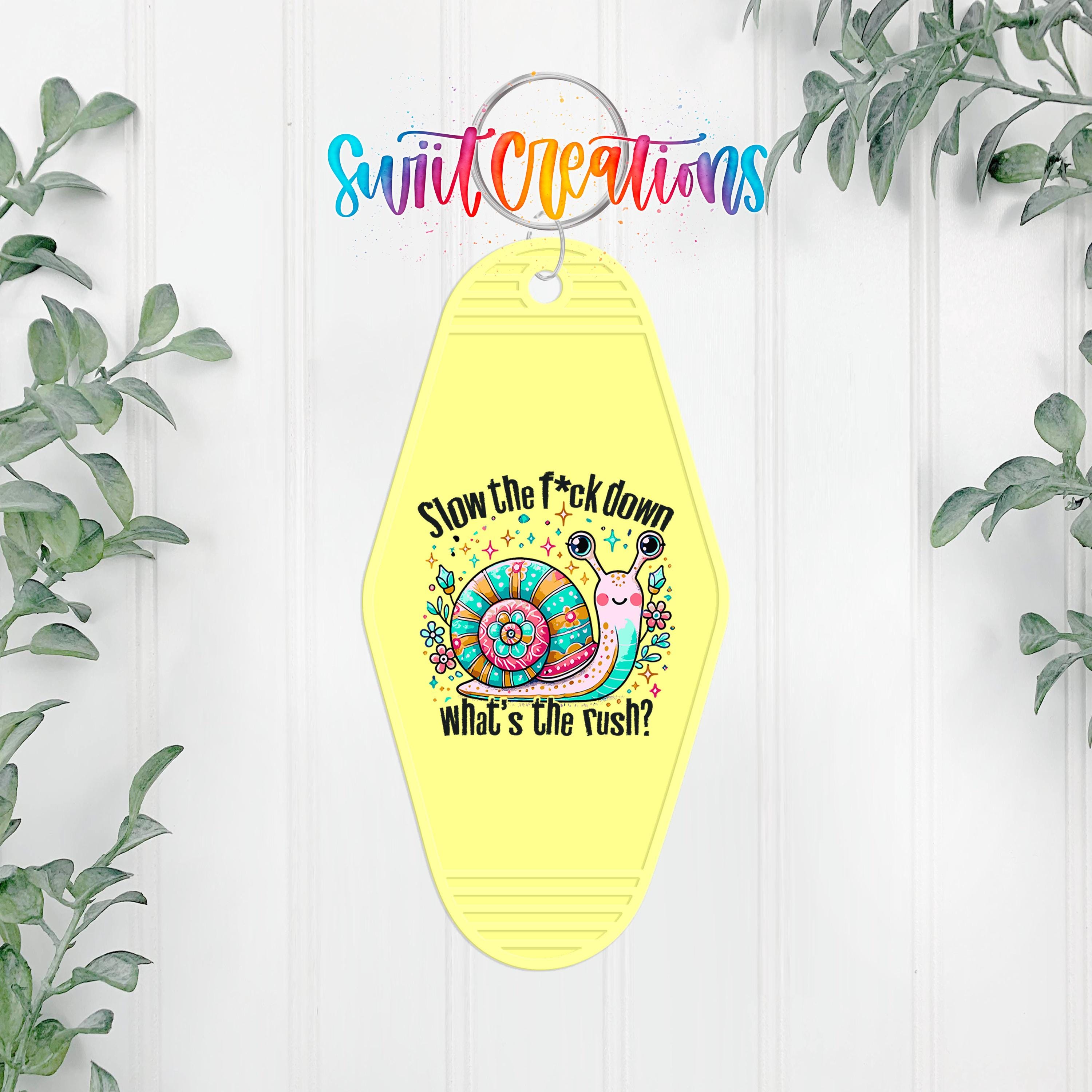 A yellow keychain with a colorful snail design and the text "Slow the fuck down what's the rush?" hanging on a wall with greenery.