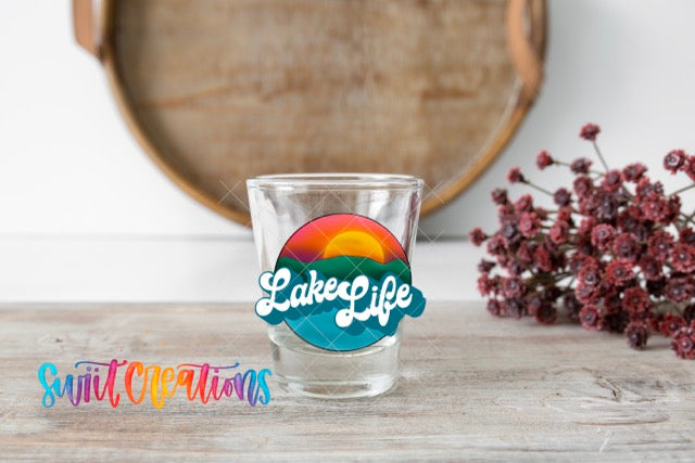 A shot glass with a colorful design and the text "LakeLife" on it, placed on a wooden surface next to a bunch of red flowers.