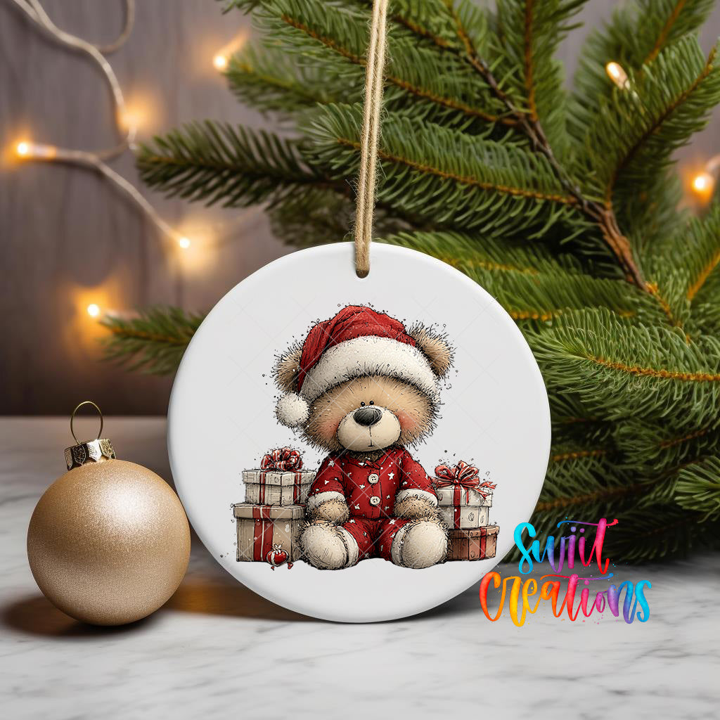 A white ceramic ornament featuring a teddy bear wearing a Santa hat and holding presents, placed on a marble surface next to a gold ornament and a Christmas tree.