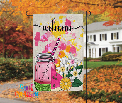 A colorful welcome sign featuring a mason jar with a straw, watermelon slices, and flowers, set against a backdrop of autumn foliage.