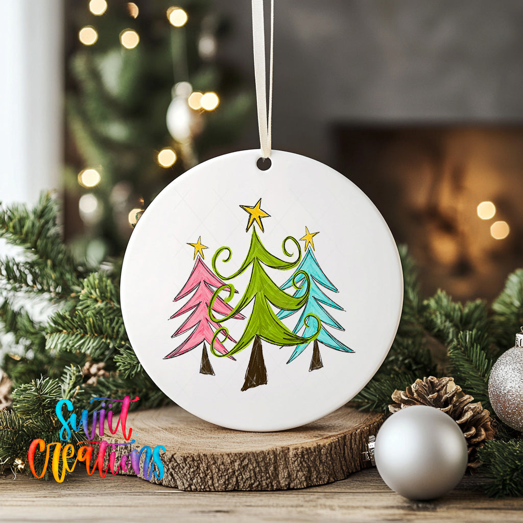 A white circular ornament with a drawing of three stylized Christmas trees in various colors hangs on a wooden surface, surrounded by pine branches and ornaments.