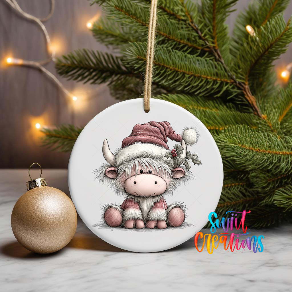 A ceramic ornament featuring a cartoon cow wearing a Santa hat, sitting on a Christmas tree branch with a gold ornament next to it.