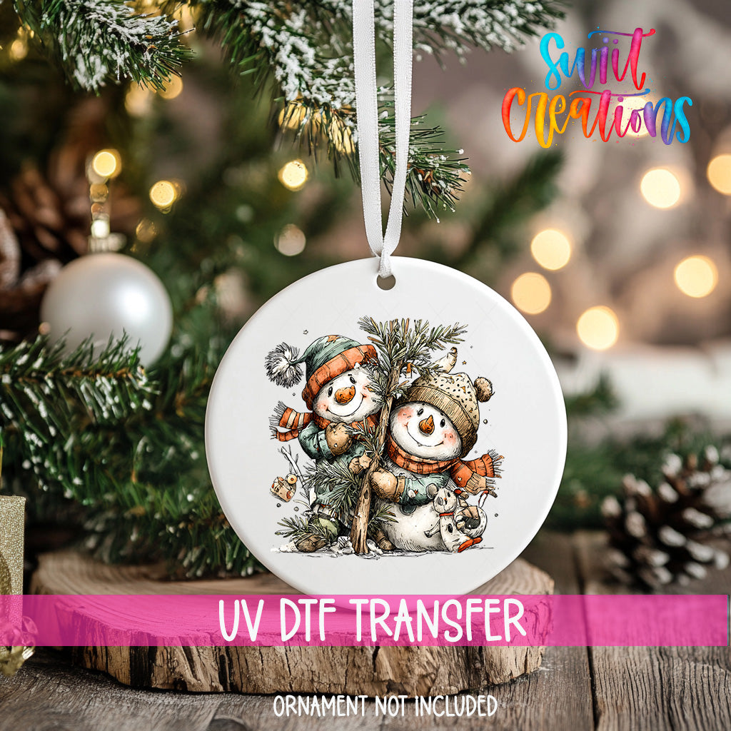 A white ceramic ornament with a snowman and pine tree design hangs on a Christmas tree.