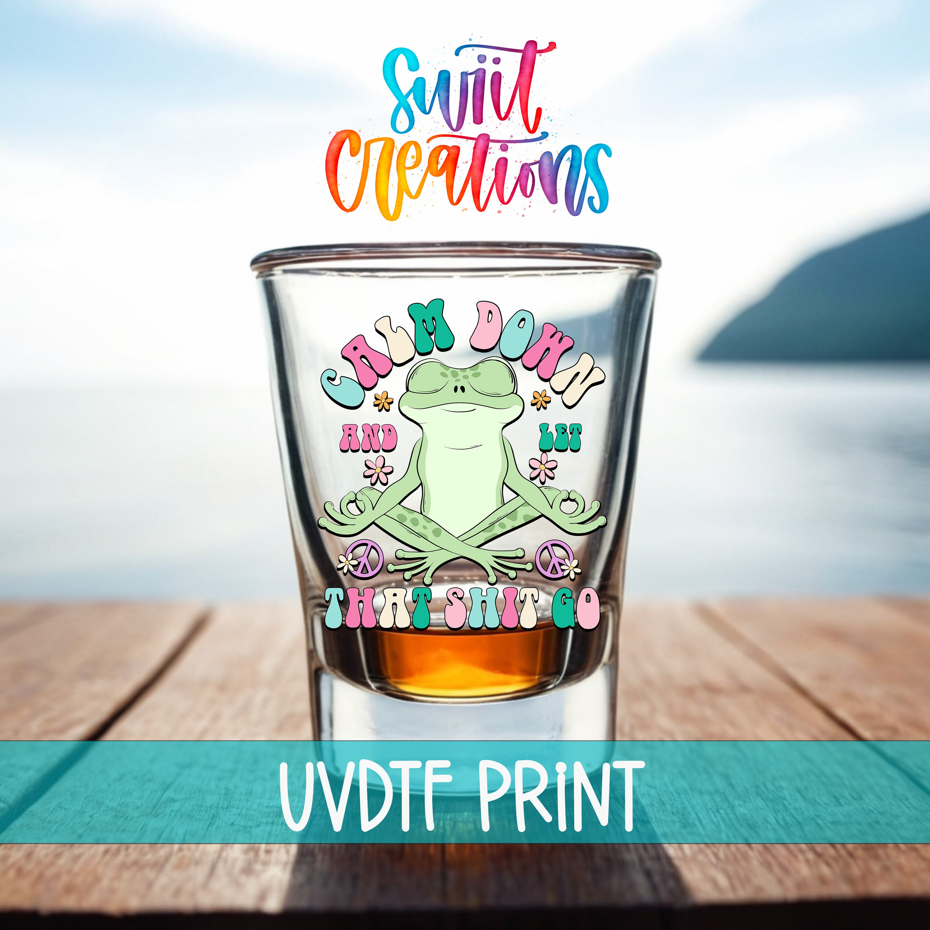 A shot glass with a frog design and text on it, placed on a wooden surface with a body of water in the background.