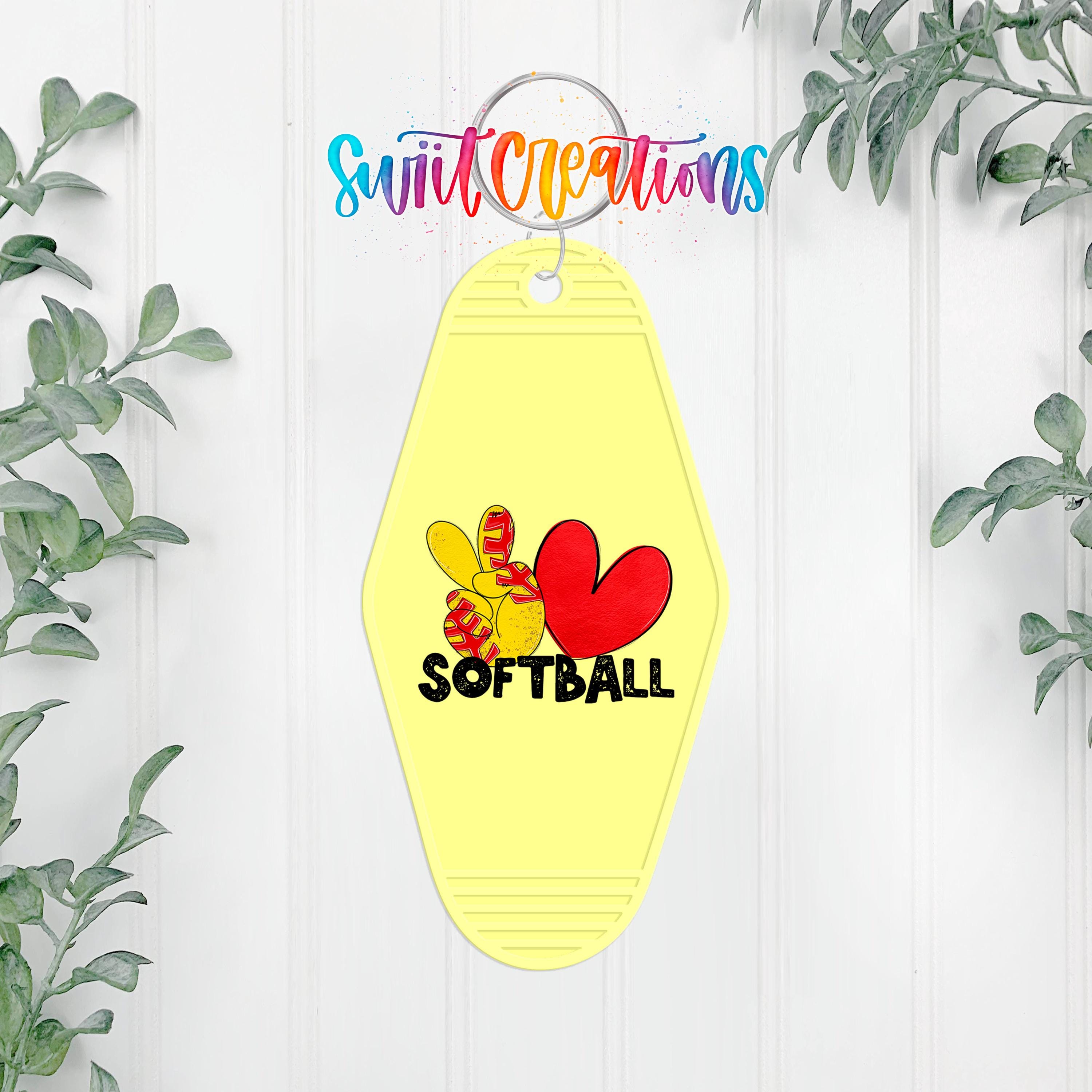 A yellow keychain with a heart and softball design, hanging on a white wall with greenery in the background.
