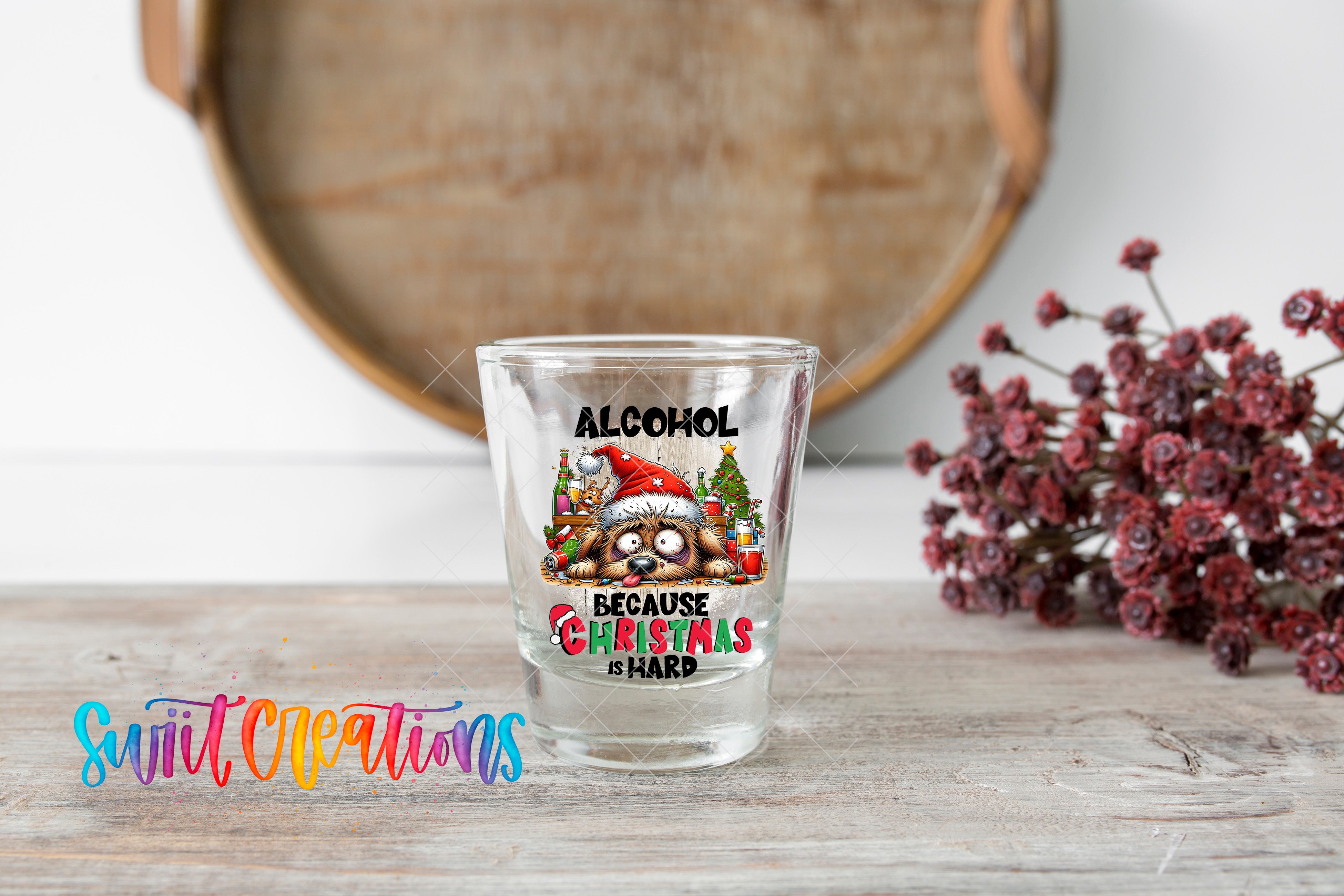 A shot glass with a humorous Christmas-themed design and text, placed on a wooden surface next to a bunch of red flowers.