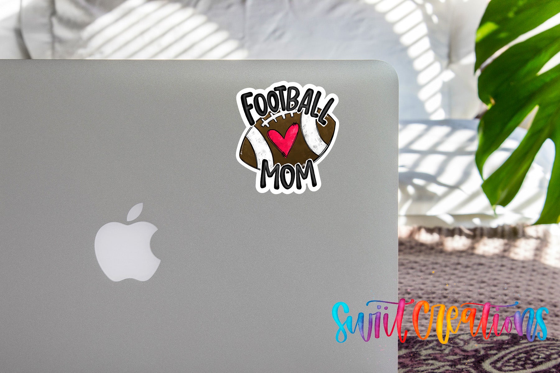 a sticker with the text "Football Mom" on the back of a silver laptop, with a plant visible in the background.