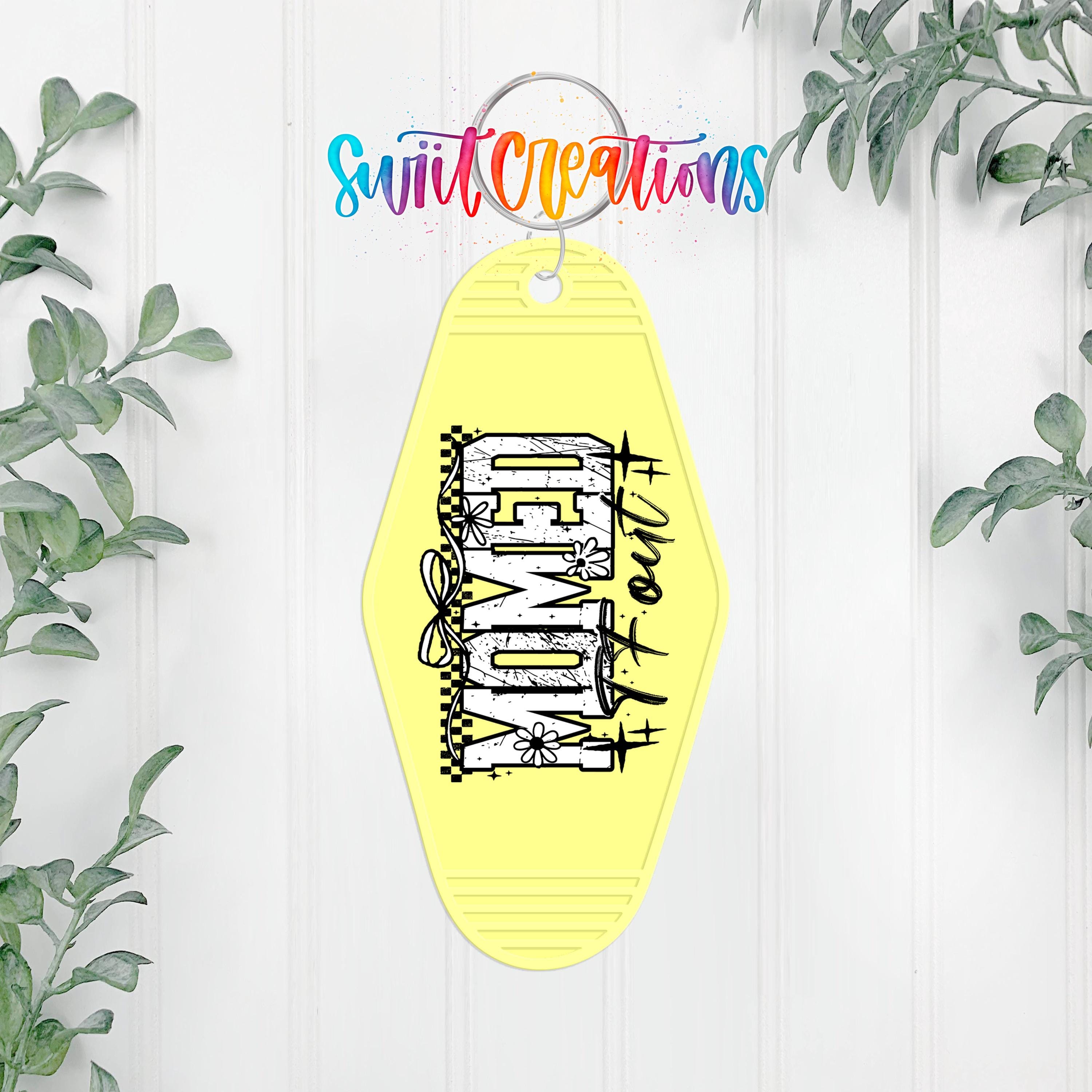 A yellow keychain with the word "DEAD" printed on it, hanging on a white wall with some greenery in the background.