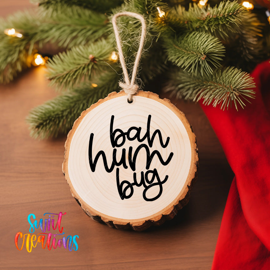 A wooden ornament with the text "bah hum bug" hanging from a tree branch, surrounded by pine branches and twinkling lights.